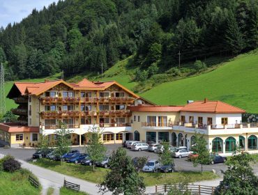 Ein großes Alpenhotel mit Balkonen, eingebettet in einen bewaldeten Hang, umgeben von parkenden Autos und Grünanlagen.