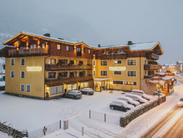 Verschneite Abendansicht des Hotels Tauernhof mit geparkten Autos und warmen Lichtern in einem Bergdorf.
