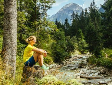 Frau in Sportkleidung sitzt auf einem Baumstumpf an einem Waldbach in der Nähe der SalzburgerLand Hotels, im Hintergrund die Berge.