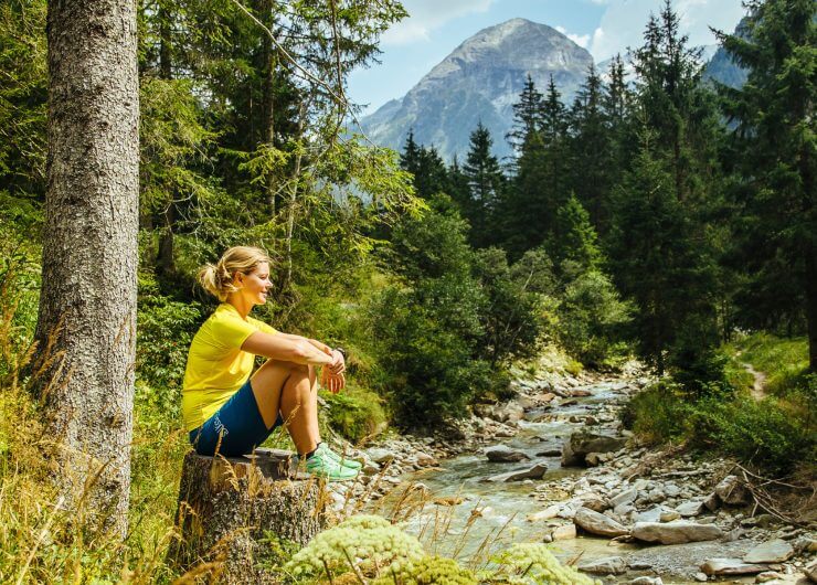 Frau in Sportkleidung sitzt auf einem Baumstumpf an einem Waldbach in der Nähe der SalzburgerLand Hotels, im Hintergrund die Berge.