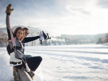 Eislaufen-(c)-Robert-Kittel Zwei Personen sitzen auf einer verschneiten Bank in Saalfelden Leogang, lächelnd mit Schlittschuhen inmitten einer Winterlandschaft.