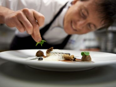 photographer Ein Koch garniert sorgfältig ein Waldrezepte-Gourmetgericht auf einem weißen Teller in einer professionellen Küche.