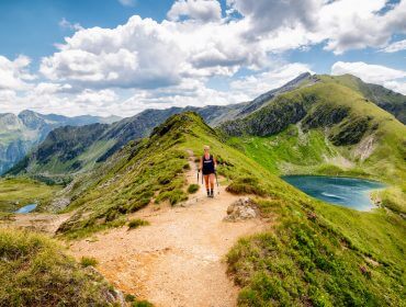 Eine Person wandert entlang eines Bergkamms im Naturwellness Lungau, mit grünen Hügeln, einem See und dramatischen Wolken im Hintergrund.