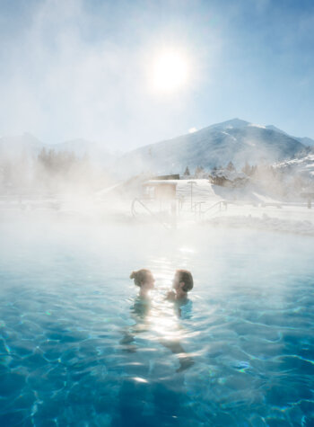 Zwei Personen entspannen sich in einem dampfenden Außenpool der Alpentherme Gastein mit schneebedeckten Bergen und strahlender Sonne im Hintergrund.