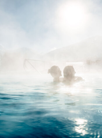 Zwei Personen in einem dampfenden Freibad der Alpentherme Gastein mit schneebedeckten Bergen und strahlender Sonne im Hintergrund.
