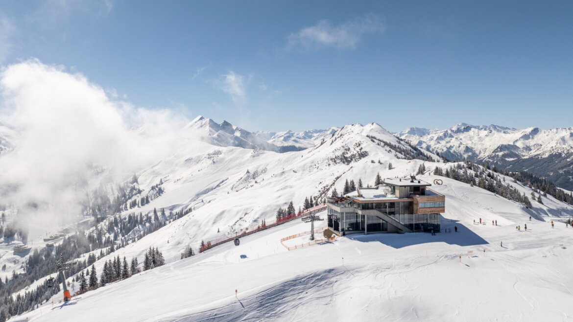 Moderne Skihütte auf einem verschneiten Berggipfel mit umliegender alpiner Landschaft unter strahlend blauem Himmel. (vergrößerte Ansicht)