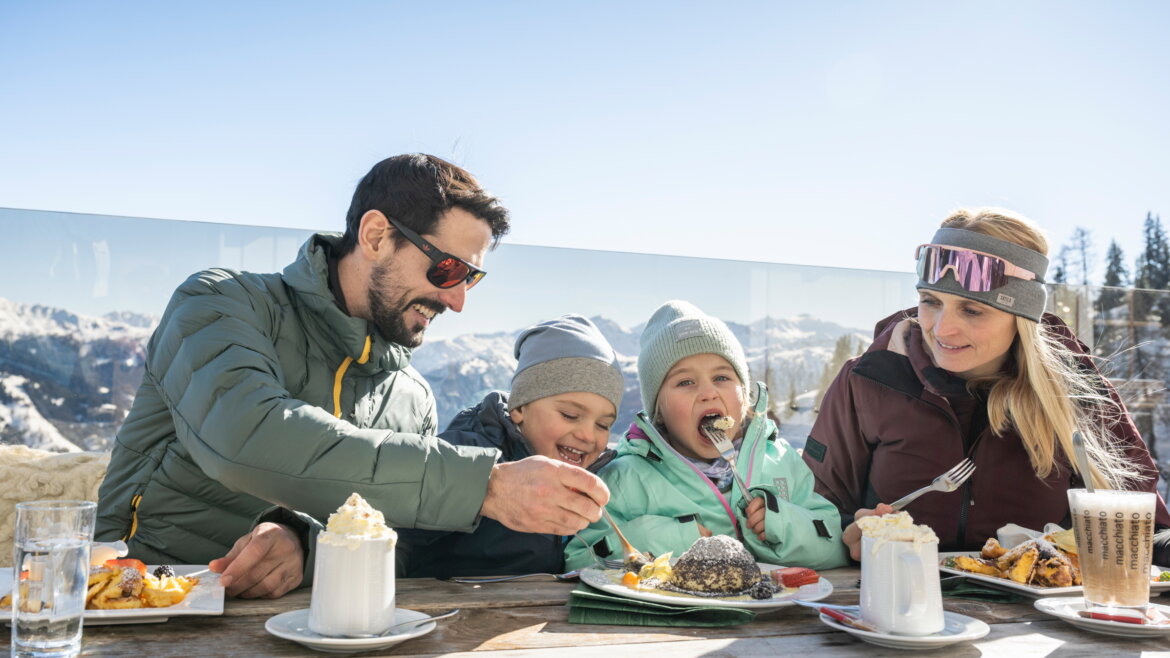 Eine Familie genießt eine Mahlzeit und heiße Getränke im Freien mit verschneiten Bergen im Hintergrund. (vergrößerte Ansicht)