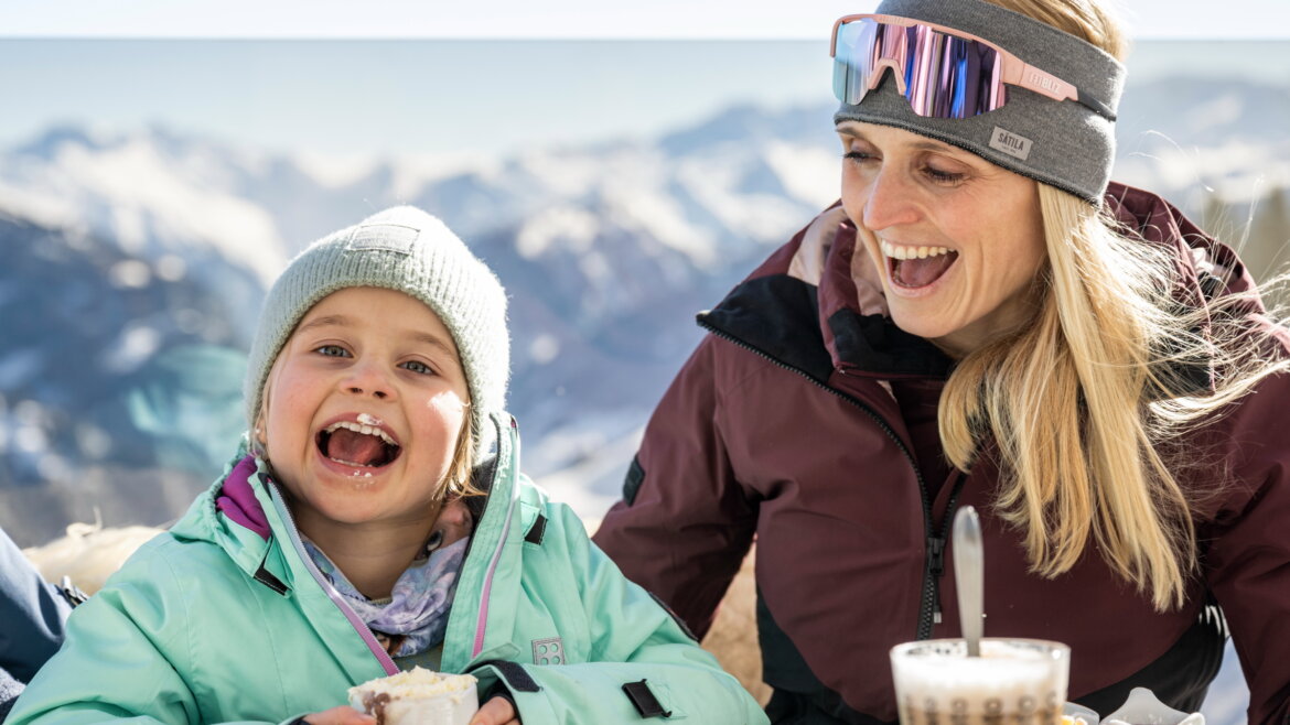 Lächelnde Frau und Kind in Winterkleidung genießen heiße Getränke und Essen im Freien mit verschneiten Bergen im Hintergrund. (vergrößerte Ansicht)