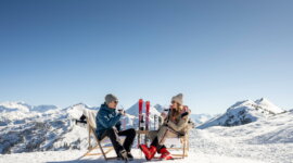 Grossarltal Ski- und Weingenuss © LorenzMasser Zwei Personen in Winterkleidung sitzen auf Stühlen im Schnee, die Berge im Rücken, und genießen einen Drink.