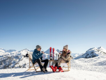 Zwei Personen in Winterkleidung sitzen auf Stühlen im Schnee, die Berge im Rücken, und genießen einen Drink.