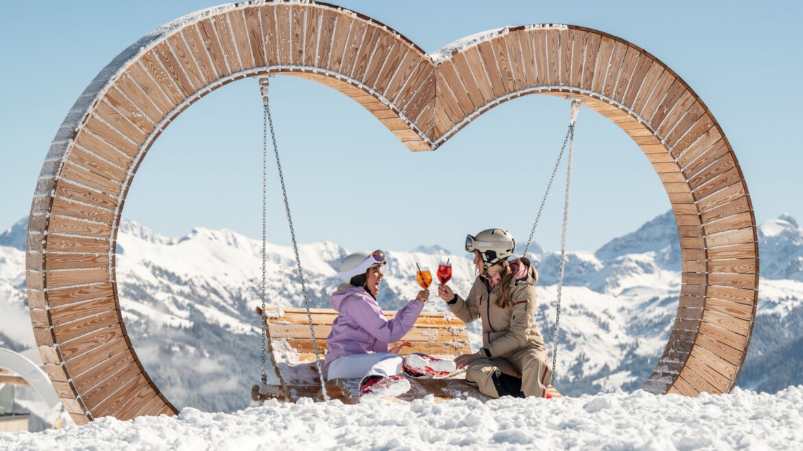 Zwei Menschen auf einer herzförmigen Schaukel in den verschneiten Bergen, die unter einem klaren blauen Himmel mit Getränken anstoßen. (vergrößerte Ansicht)