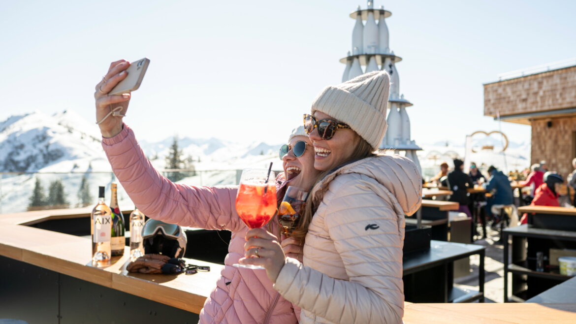 Zwei Frauen in Winterjacken machen ein Selfie und stoßen in einer sonnigen Bar in einem Skigebiet mit Getränken an. (vergrößerte Ansicht)