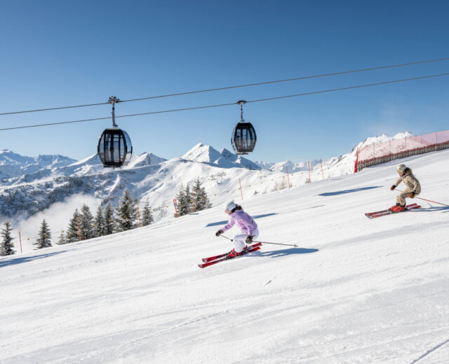 Grossarltal Skifahren © Lorenz Masser Zwei Skifahrer fahren einen verschneiten Berghang unter Skiliftgondeln hinunter, mit schneebedeckten Gipfeln im Hintergrund.