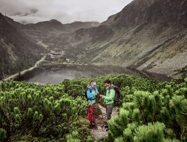 Zwei Wanderer auf einem schmalen Pfad oberhalb eines Sees, genießen die walderlebnisse uttendorf inmitten üppiger Berge und wolkenverhangenem Himmel.