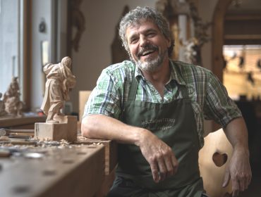 Ein lächelnder Holzschnitzer in einer Schürze sitzt in einer Holzwerkstatt neben einer Holzskulptur und Schnitzwerkzeugen.