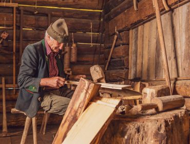 Älterer Mann in rustikaler österreichischer Kleidung schnitzt Holz in einer Hüttenwerkstatt im Walderlebnis Saalachtal.