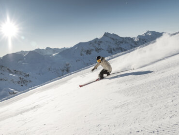Obertauern Skifahren (c)Mirja Geh,Skiing,Austria Ein Skifahrer fährt einen sonnigen, schneebedeckten Berghang hinunter, mit fernen Gipfeln im Hintergrund.
