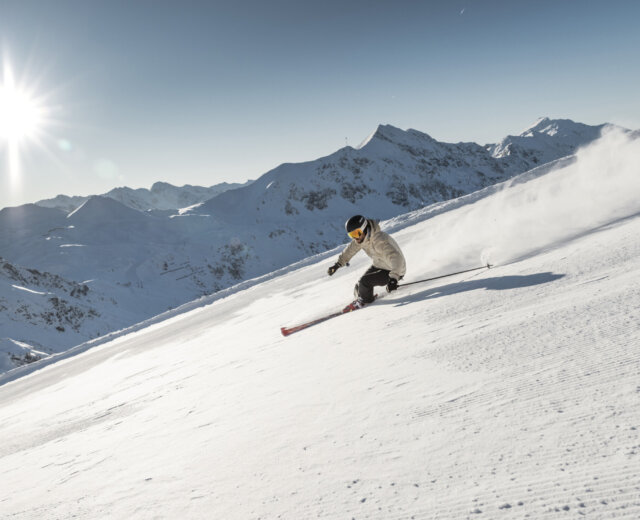 Obertauern Skifahren (c)Mirja Geh,Skiing,Austria Ein Skifahrer fährt einen sonnigen, schneebedeckten Berghang hinunter, mit fernen Gipfeln im Hintergrund.