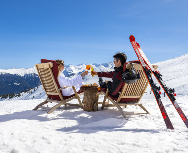 Chillout Skifahren (C) Gasteinertal Tourismus GmbH, Max Steinbauer Zwei Personen in Skikleidung stoßen in Liegestühlen auf einem verschneiten Berg mit Skiern und schneebedeckten Gipfeln im Hintergrund an.