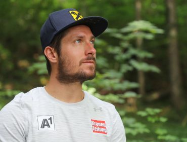 Ein bärtiger Mann mit Mütze und Sporthemd blickt auf, der Marcel Hirscher ähnelt, während er sich in einem Waldgebiet befindet.