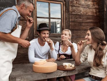 Vier Personen in traditioneller Kleidung genießen Käse an einem rustikalen Tisch vor einer Berghütte in Saalfelden Leogang.