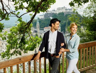 RS2289_pärchen-mönchsberg_2 Ein lächelndes Paar geht an einem Holzzaun, Stadtwäldern und einer malerischen Stadt mit einer Burg im Hintergrund vorbei.