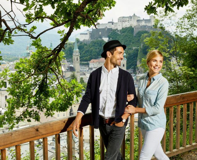 RS2289_pärchen-mönchsberg_2 Ein lächelndes Paar geht an einem Holzzaun, Stadtwäldern und einer malerischen Stadt mit einer Burg im Hintergrund vorbei.