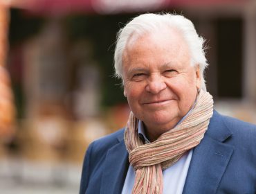 Älterer Mann mit weißem Haar, der einen blauen Blazer und einen Schal trägt, lächelt im Freien vor einem unscharfen Hintergrund.