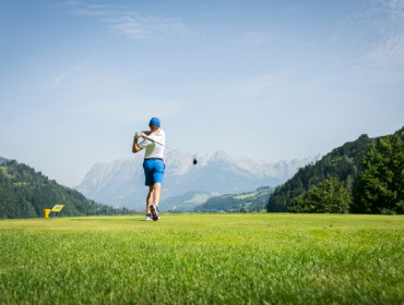 Ein Golfer schwingt auf einem grünen Platz mit Bergen und blauem Himmel im Hintergrund.