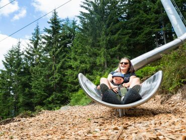 Eine Frau und ein Kind lächeln, während sie eine glänzende Metallrutsche hinunterfahren, die von Bäumen und Holzspänen umgeben ist.