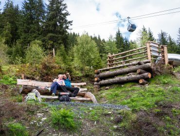 Ein Paar sitzt auf einer Holzbank in einem Waldgebiet, mit einer Seilbahn und einer Rutsche im Hintergrund.