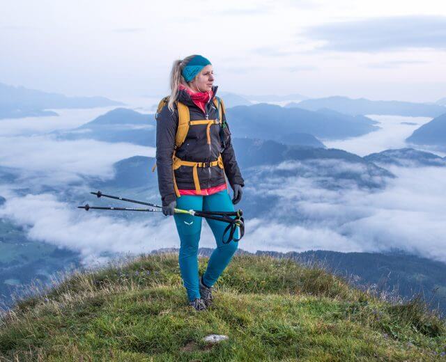 Frau steht mit Wanderstöcken auf einem Berggipfel und blickt auf Wolken und ferne Berge bei Sonnenaufgang.