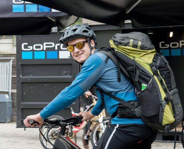 Ein lächelnder Radfahrer mit einem großen Rucksack und Helm posiert auf einem Fahrrad vor einem GoPro-Stand.