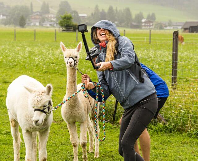 Eine Frau in einer Regenjacke macht ein Selfie mit zwei Alpakas auf einer Wiese, lächelt und hält ihre Leinen.
