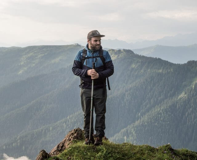 Mann mit Wanderstöcken steht auf einem grasbewachsenen Berggipfel mit bewaldeten Hügeln und bewölktem Himmel im Hintergrund.