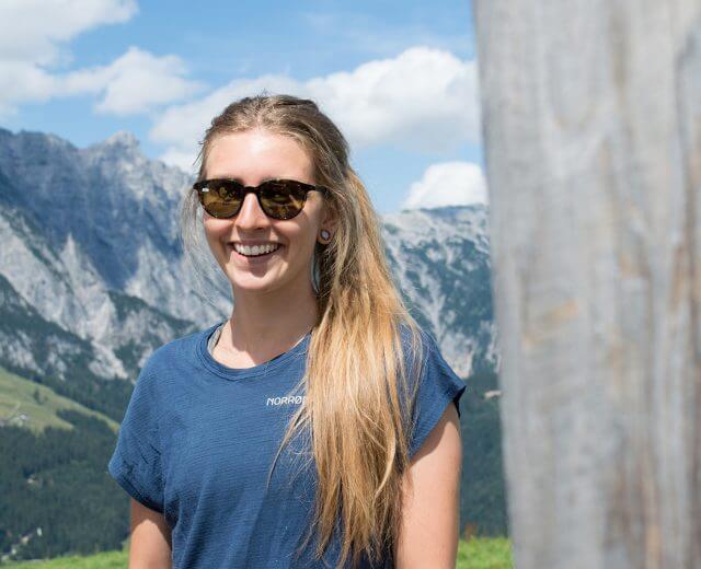 Lächelnde Frau mit Sonnenbrille steht im Freien mit Bergen und blauem Himmel im Hintergrund.