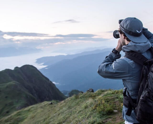 Eine Person fotografiert die Berge bei Sonnenaufgang mit Rucksack und Mütze auf einem grasbewachsenen Bergrücken.