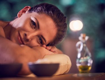 Eine Frau entspannt sich auf einer Massageliege in einem Day Spa und stützt ihren Kopf auf ihre Arme, während Schalen und Öl in der Nähe stehen.