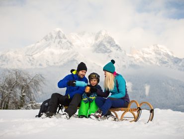 Eine Familie in Winterkleidung sitzt im Schnee in Saalfelden-Leogang und genießt ein Getränk am Schlitten mit Blick auf die Berge.