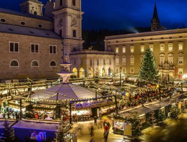 Ein festlicher Weihnachtsmarkt mit Lichtern, einem großen Baum und vielen Menschen auf einem historischen Stadtplatz bei Nacht.