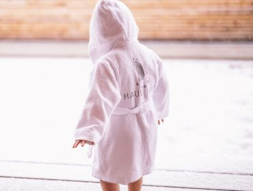 Ein kleines Kind in einem weißen Kapuzenmantel läuft barfuß auf einer verschneiten Terrasse entlang des BioParadies Weges Bad Gastein.