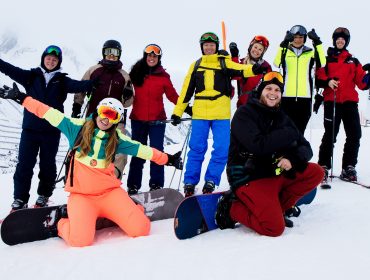 Eine Gruppe lächelnder Menschen in bunter Winterkleidung posiert mit Snowboards und Skiern in den verschneiten Bergen von Obertauern.