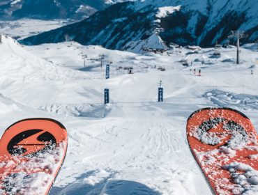 Blick eines Skifahrers auf eine verschneite Skipiste mit Bergen und Skiliften im Hintergrund.