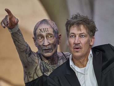 Ein tätowierter Jedermann-Schauspieler zeigt auf einen Mann im Anzug; beide haben einen dramatischen Gesichtsausdruck.