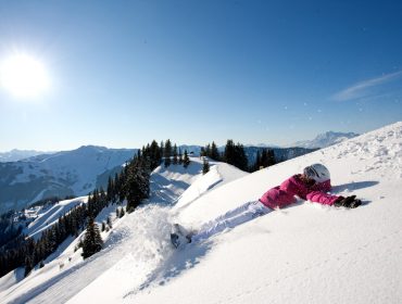 Eine Person in rosafarbener Schneekleidung fällt spielerisch in den Tiefschnee auf einer sonnigen Piste der alpinen Gesundheitsregion.