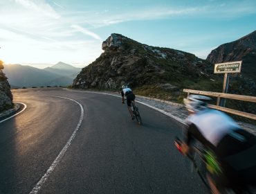 Grossglockner_06(c)WOMMedien_StefanSchopf_SalzburgerLand Zwei Radfahrer rasen bei Sonnenuntergang auf einer kurvenreichen Straße bergab und rasen wie Glocknerkönige zwischen felsigen Klippen hindurch.
