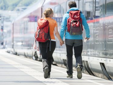 Ein Paar mit Rucksäcken hält sich auf einem Tennengauer Bahnsteig neben einem modernen Zug an den Händen.