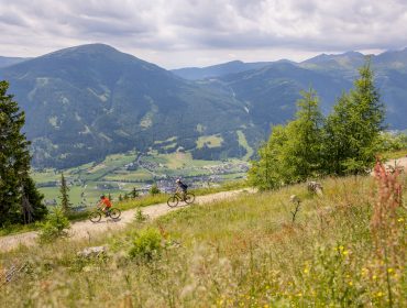 Mountainbike Großeck Speierck (2) Zwei Radfahrer fahren auf einem Bergweg im Lungau mit einem malerischen Tal und Bergen im Hintergrund.