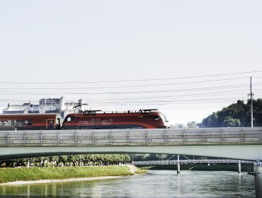 Railjet_HOR_7625_c_OEBB_PhilippHorak Ein roter Railjet-Zug überquert eine Brücke im Tennengau, mit einem Fluss, Grünflächen und einem Kreuzfahrtschiff im Hintergrund.