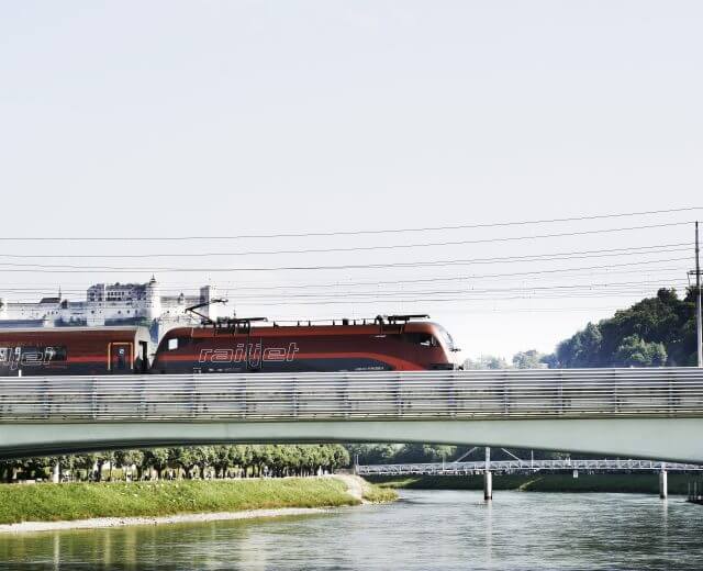 Railjet_HOR_7625_c_OEBB_PhilippHorak Ein roter Railjet-Zug überquert eine Brücke im Tennengau, mit einem Fluss, Grünflächen und einem Kreuzfahrtschiff im Hintergrund.